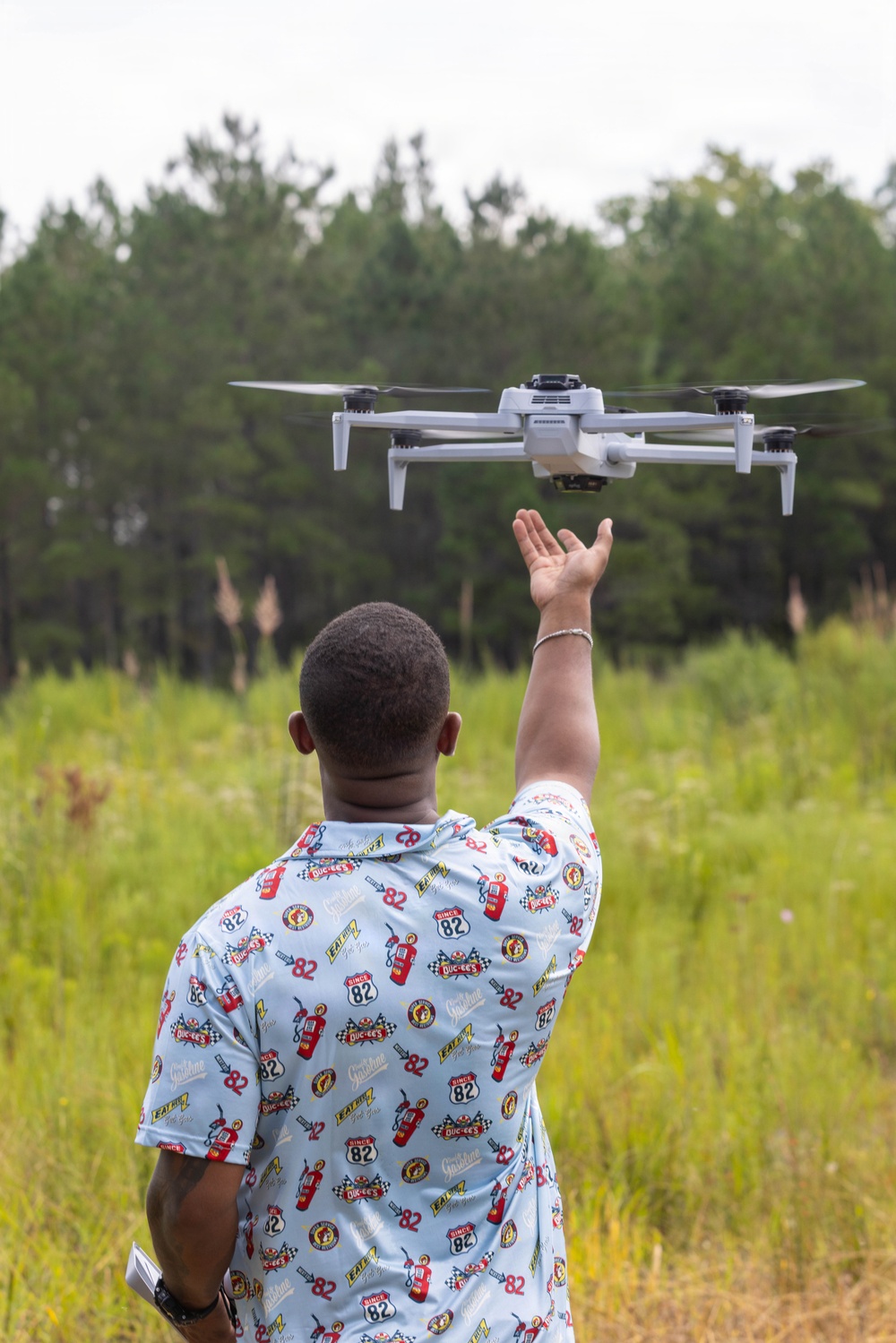 A person is standing in an open grassy field reaching up toward a hovering drone with one hand. The individual is wearing a light blue short-sleeve shirt covered in Buc-ee's branded graphics and the number 82. The setting appears to be a rural or semi-wild outdoor area with tall grass and a tree line in the background.