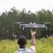 A person is standing in an open grassy field reaching up toward a hovering drone with one hand. The individual is wearing a light blue short-sleeve shirt covered in Buc-ee's branded graphics and the number 82. The setting appears to be a rural or semi-wild outdoor area with tall grass and a tree line in the background.