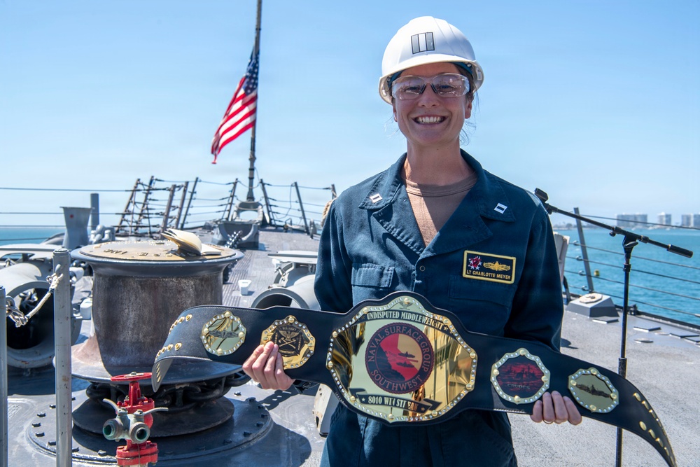 A U.S. Navy Lieutenant named Charlotte Meyer, wearing a white hard hat with captain's bars insignia and blue coveralls, stands on the deck of a naval vessel holding a championship-style belt from Naval Surface Group Southwest. She is smiling broadly while displaying the ornate gold and black belt, which reads 'Undisputed Middleweight Naval Surface Group Southwest.' An American flag is visible in the background against a clear blue sky.