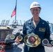 A U.S. Navy Lieutenant named Charlotte Meyer, wearing a white hard hat with captain's bars insignia and blue coveralls, stands on the deck of a naval vessel holding a championship-style belt from Naval Surface Group Southwest. She is smiling broadly while displaying the ornate gold and black belt, which reads 'Undisputed Middleweight Naval Surface Group Southwest.' An American flag is visible in the background against a clear blue sky.