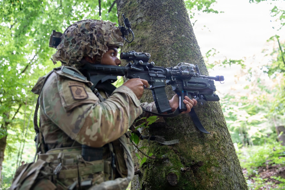 A soldier in full camouflage gear is positioned behind a large tree trunk in a forested environment, aiming a rifle with an optical scope and accessories. The soldier is wearing a multicam uniform with patches, a camouflage helmet cover, and tactical equipment. The setting appears to be a military training exercise or field operation in a deciduous woodland.