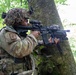 A soldier in full camouflage gear is positioned behind a large tree trunk in a forested environment, aiming a rifle with an optical scope and accessories. The soldier is wearing a multicam uniform with patches, a camouflage helmet cover, and tactical equipment. The setting appears to be a military training exercise or field operation in a deciduous woodland.