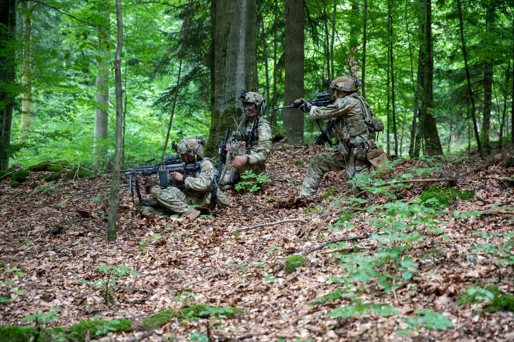 Three soldiers in military camouflage uniforms are positioned in a forested area, appearing to conduct a tactical field exercise or training operation. They are arranged in a small unit formation on a leaf-covered hillside among tall trees. The soldiers are armed and appear to be in active tactical postures, suggesting a coordinated maneuver or defensive position.