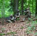 Three soldiers in military camouflage uniforms are positioned in a forested area, appearing to conduct a tactical field exercise or training operation. They are arranged in a small unit formation on a leaf-covered hillside among tall trees. The soldiers are armed and appear to be in active tactical postures, suggesting a coordinated maneuver or defensive position.