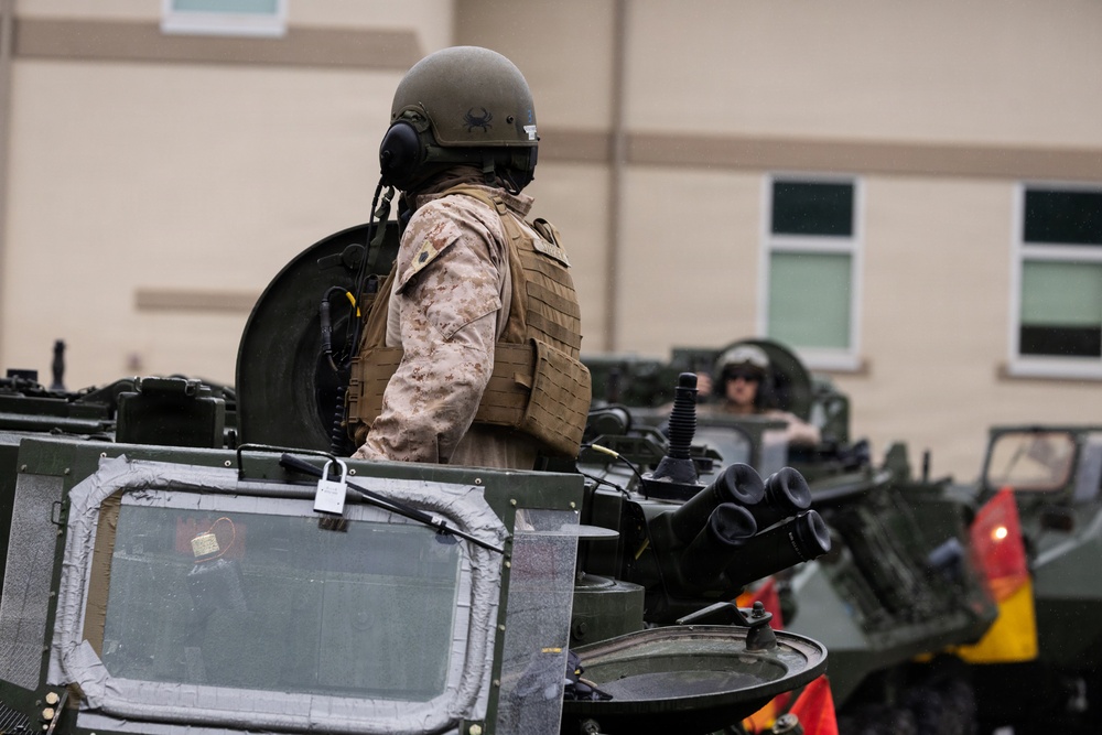 A military personnel wearing desert camouflage (MARPAT pattern) uniform, tactical vest, and combat helmet is positioned in the hatch of an armored military vehicle. At least two soldiers and multiple armored vehicles are visible, suggesting a convoy formation or staging area near a building. The scene appears to be a military base or installation.