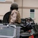 A military personnel wearing desert camouflage (MARPAT pattern) uniform, tactical vest, and combat helmet is positioned in the hatch of an armored military vehicle. At least two soldiers and multiple armored vehicles are visible, suggesting a convoy formation or staging area near a building. The scene appears to be a military base or installation.