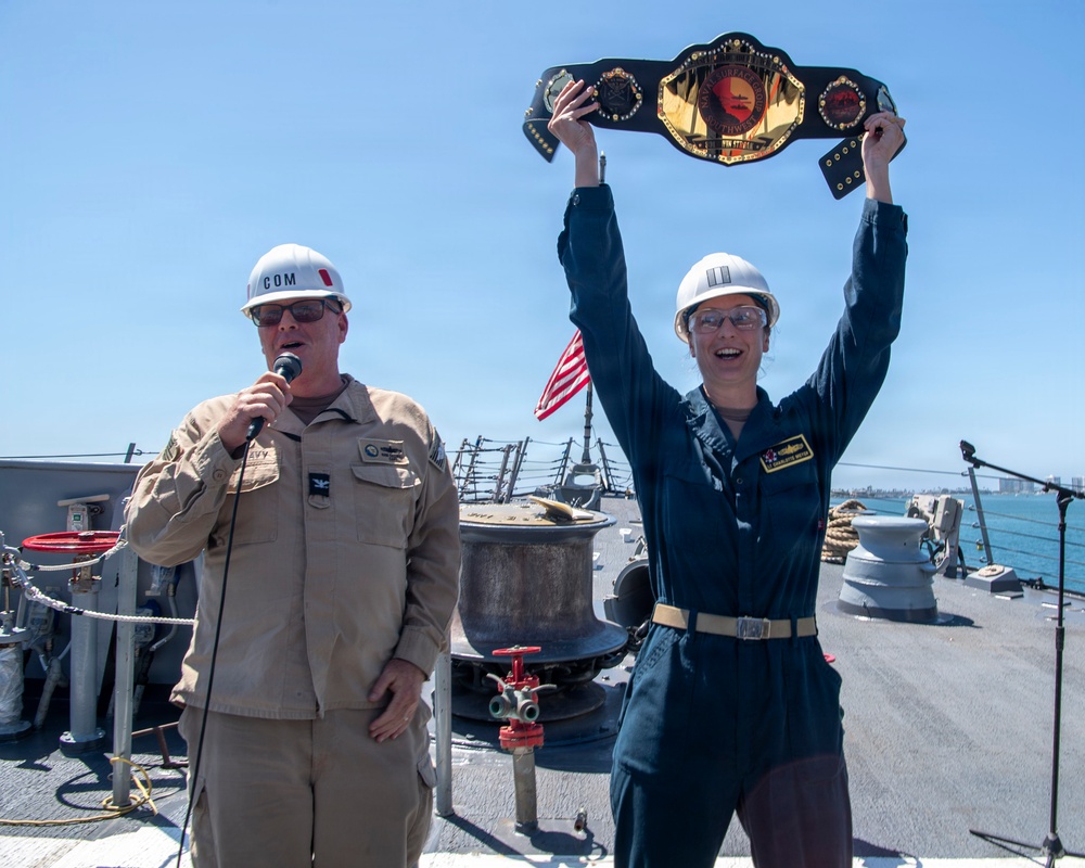 Two U.S. Navy personnel are standing on the deck of a naval vessel. One person in tan coveralls holds a microphone and speaks, while another in dark blue coveralls triumphantly raises a large championship-style belt overhead. The scene appears to be an award or recognition ceremony conducted on the ship's deck.