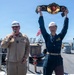 Two U.S. Navy personnel are standing on the deck of a naval vessel. One person in tan coveralls holds a microphone and speaks, while another in dark blue coveralls triumphantly raises a large championship-style belt overhead. The scene appears to be an award or recognition ceremony conducted on the ship's deck.