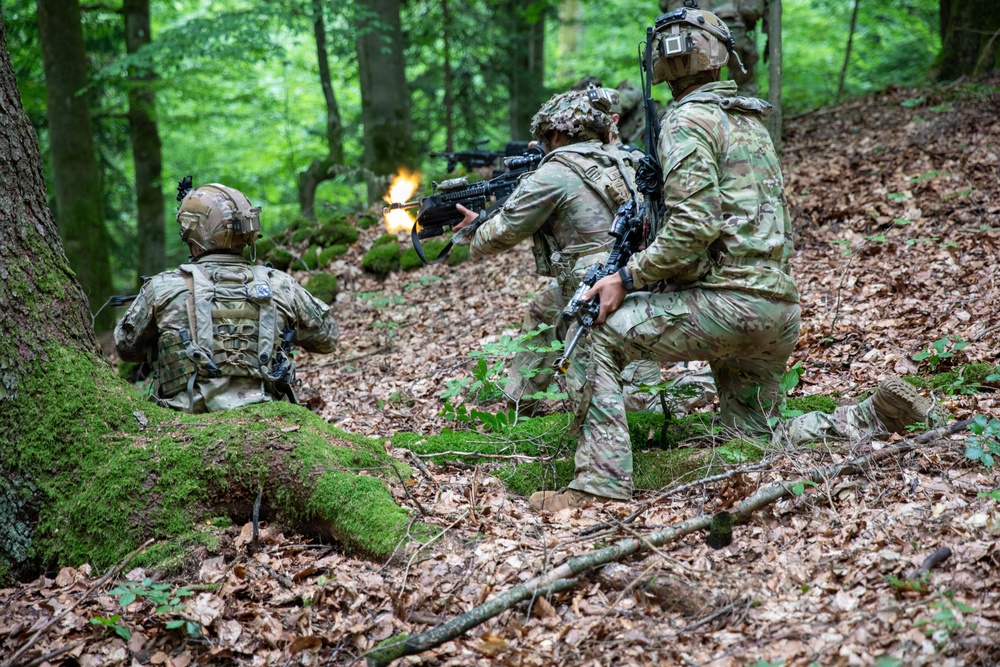 Three soldiers in camouflage uniforms and tactical gear are conducting a woodland combat exercise on a leaf-covered hillside. One soldier is actively firing a weapon with a visible muzzle flash, while the others maintain tactical positions. The setting appears to be a deciduous forest with moss-covered logs and green foliage.