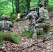 Three soldiers in camouflage uniforms and tactical gear are conducting a woodland combat exercise on a leaf-covered hillside. One soldier is actively firing a weapon with a visible muzzle flash, while the others maintain tactical positions. The setting appears to be a deciduous forest with moss-covered logs and green foliage.