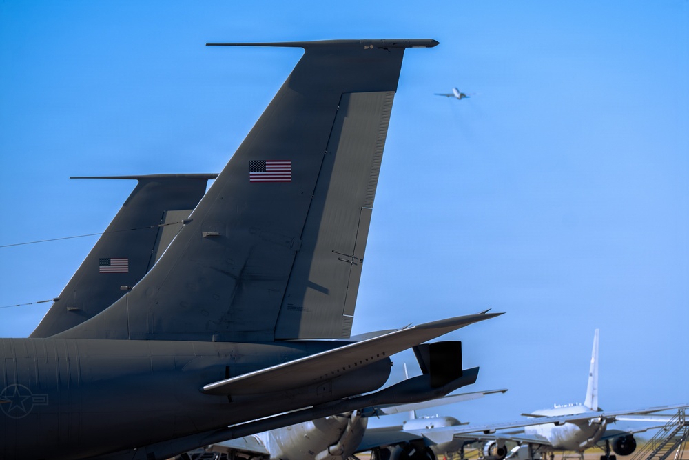 The image shows multiple large military aircraft parked on a flight line, with their distinctive tail sections prominently displayed in the foreground. American flags are visible on the tail fins, identifying these as United States Air Force aircraft, likely KC-135 Stratotankers or similar large military jets. A fourth aircraft is visible in the background taking off or flying at low altitude against a clear blue sky.