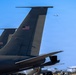The image shows multiple large military aircraft parked on a flight line, with their distinctive tail sections prominently displayed in the foreground. American flags are visible on the tail fins, identifying these as United States Air Force aircraft, likely KC-135 Stratotankers or similar large military jets. A fourth aircraft is visible in the background taking off or flying at low altitude against a clear blue sky.