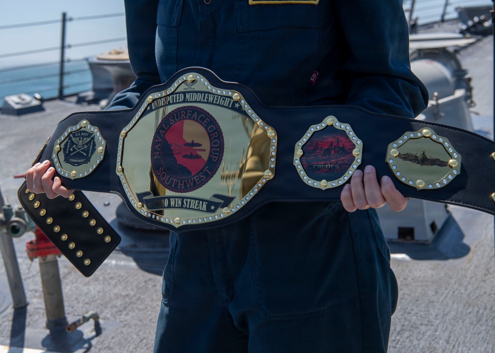 A person in U.S. Navy dark blue coveralls holds a large championship-style belt on what appears to be the deck of a naval vessel. The belt is customized for Naval Surface Group Southwest and reads 'Undisputed Middleweight' with a win streak inscription. The belt features naval imagery and insignia, styled after professional wrestling championship belts.