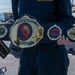A person in U.S. Navy dark blue coveralls holds a large championship-style belt on what appears to be the deck of a naval vessel. The belt is customized for Naval Surface Group Southwest and reads 'Undisputed Middleweight' with a win streak inscription. The belt features naval imagery and insignia, styled after professional wrestling championship belts.