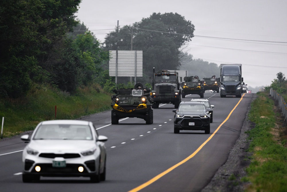 A military convoy is traveling along a two-lane highway in what appears to be a rural or semi-rural area under overcast skies. The convoy consists of multiple military vehicles with distinctive yellow markings, interspersed with civilian vehicles traveling in the same direction. The scene suggests a military movement or transport operation on a public road.