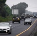 A military convoy is traveling along a two-lane highway in what appears to be a rural or semi-rural area under overcast skies. The convoy consists of multiple military vehicles with distinctive yellow markings, interspersed with civilian vehicles traveling in the same direction. The scene suggests a military movement or transport operation on a public road.