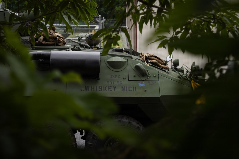 A United States Marine Corps (USMC) armored vehicle, serial number 531356, named 'WHISKEY NICK' is partially visible through tree foliage. The olive-drab painted vehicle appears to be an LAV (Light Armored Vehicle) or similar wheeled armored personnel carrier. The vehicle is positioned near a building with a chain-link fence visible in the background.