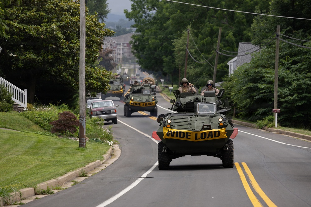 A convoy of military armored vehicles is traveling along a two-lane suburban road lined with trees and residential properties. The lead vehicle and at least two others behind it display yellow 'WIDE LOAD' banners across their fronts, indicating an official road movement. Personnel in military helmets and gear are visible riding in the open turret hatches of the vehicles.