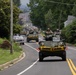 A convoy of military armored vehicles is traveling along a two-lane suburban road lined with trees and residential properties. The lead vehicle and at least two others behind it display yellow 'WIDE LOAD' banners across their fronts, indicating an official road movement. Personnel in military helmets and gear are visible riding in the open turret hatches of the vehicles.