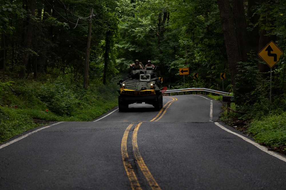 An armored military vehicle is traveling down a narrow two-lane road through a densely wooded area. Two personnel are visible in open hatches on top of the vehicle, wearing helmets and military gear. The road features double yellow center lines and road signs indicating curves ahead.