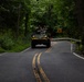 An armored military vehicle is traveling down a narrow two-lane road through a densely wooded area. Two personnel are visible in open hatches on top of the vehicle, wearing helmets and military gear. The road features double yellow center lines and road signs indicating curves ahead.