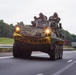 A military armored personnel carrier, likely an LAV or AAV (Amphibious Assault Vehicle), is traveling on a public highway with a yellow 'WIDE LOAD' banner across its front. At least two soldiers in helmets are visible in open hatches on top of the vehicle. A second military vehicle is visible in the background on the same road.