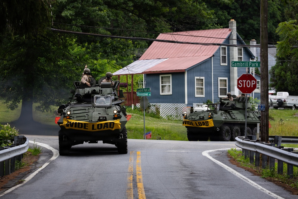 Two military armored vehicles, likely LAV-25 or similar wheeled armored personnel carriers, are traveling along a rural two-lane road in what appears to be a small American town. Both vehicles display yellow 'WIDE LOAD' banners on their fronts and have uniformed soldiers visible in their turrets. The scene is set at an intersection marked 'Gambrill Park' and 'Shookstown Rd' with a stop sign visible.