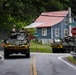 Two military armored vehicles, likely LAV-25 or similar wheeled armored personnel carriers, are traveling along a rural two-lane road in what appears to be a small American town. Both vehicles display yellow 'WIDE LOAD' banners on their fronts and have uniformed soldiers visible in their turrets. The scene is set at an intersection marked 'Gambrill Park' and 'Shookstown Rd' with a stop sign visible.