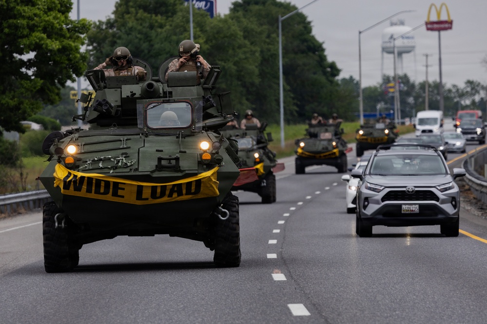 A convoy of military wheeled armored vehicles, likely LAV-25 or similar 8-wheeled armored personnel carriers, travels along a public highway in what appears to be the United States. The lead vehicle displays a yellow 'WIDE LOAD' banner across its front, indicating a permitted oversized vehicle movement on a civilian road. Civilian traffic, including a Toyota RAV4, shares the road alongside the convoy.
