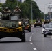 A convoy of military wheeled armored vehicles, likely LAV-25 or similar 8-wheeled armored personnel carriers, travels along a public highway in what appears to be the United States. The lead vehicle displays a yellow 'WIDE LOAD' banner across its front, indicating a permitted oversized vehicle movement on a civilian road. Civilian traffic, including a Toyota RAV4, shares the road alongside the convoy.