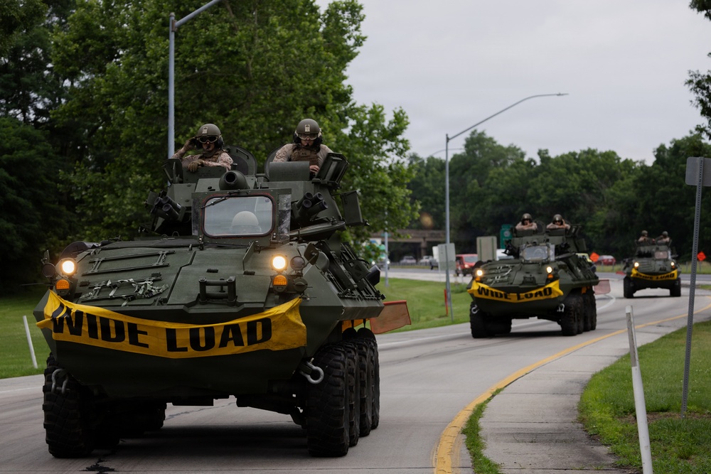 A convoy of military armored wheeled vehicles, likely LAV-25 or similar light armored vehicles, travels along a public road. The vehicles are marked with yellow 'WIDE LOAD' banners, indicating an authorized road movement. Military personnel in helmets and combat gear are visible in the open hatches of each vehicle.