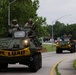 A convoy of military armored wheeled vehicles, likely LAV-25 or similar light armored vehicles, travels along a public road. The vehicles are marked with yellow 'WIDE LOAD' banners, indicating an authorized road movement. Military personnel in helmets and combat gear are visible in the open hatches of each vehicle.