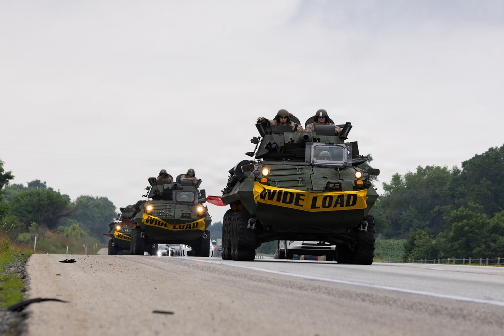 Two military armored wheeled vehicles, likely LAV-25s or similar 8-wheeled armored personnel carriers, are traveling in convoy on a public highway. Each vehicle displays a yellow 'WIDE LOAD' banner across its front, indicating compliance with civilian road transport regulations. Military personnel in helmets and combat gear are visible riding in the open hatches of both vehicles.
