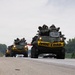 Two military armored wheeled vehicles, likely LAV-25s or similar 8-wheeled armored personnel carriers, are traveling in convoy on a public highway. Each vehicle displays a yellow 'WIDE LOAD' banner across its front, indicating compliance with civilian road transport regulations. Military personnel in helmets and combat gear are visible riding in the open hatches of both vehicles.