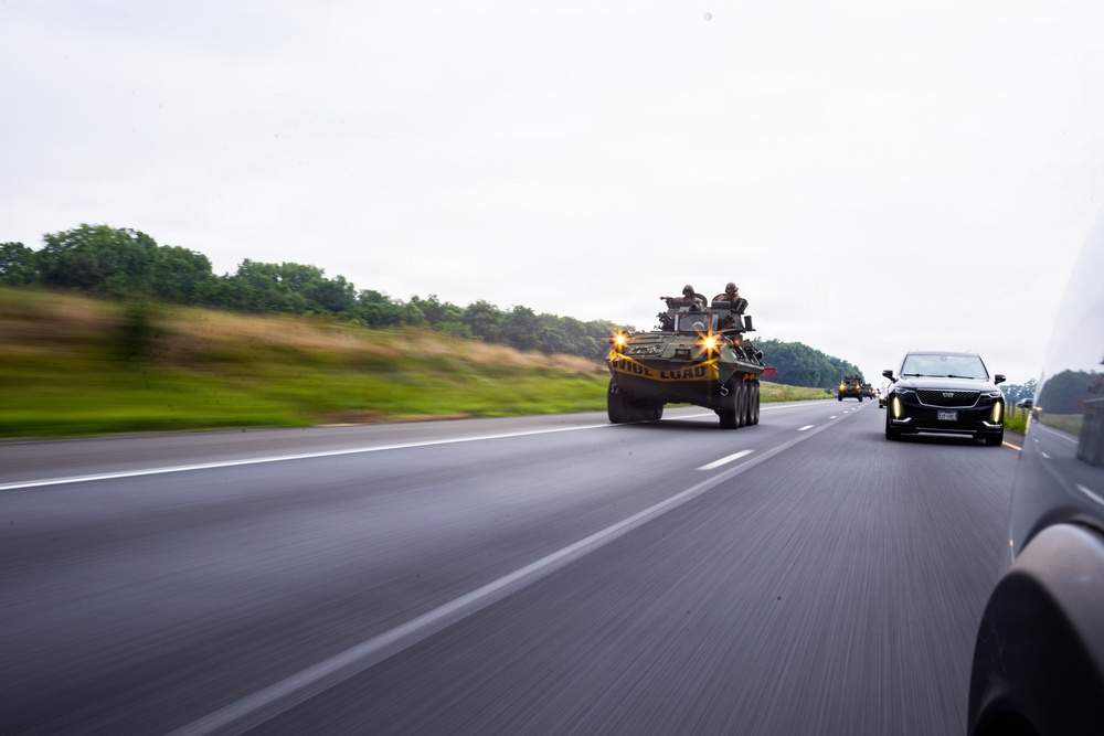 A military armored vehicle bearing a 'WIDE LOAD' banner is being transported on a multi-lane highway, accompanied by personnel riding on top of the vehicle. The convoy is moving alongside civilian traffic, including a dark-colored Cadillac SUV visible in the adjacent lane. The image was taken from a moving vehicle, producing motion blur on the road surface and background vegetation.