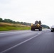 A military armored vehicle bearing a 'WIDE LOAD' banner is being transported on a multi-lane highway, accompanied by personnel riding on top of the vehicle. The convoy is moving alongside civilian traffic, including a dark-colored Cadillac SUV visible in the adjacent lane. The image was taken from a moving vehicle, producing motion blur on the road surface and background vegetation.