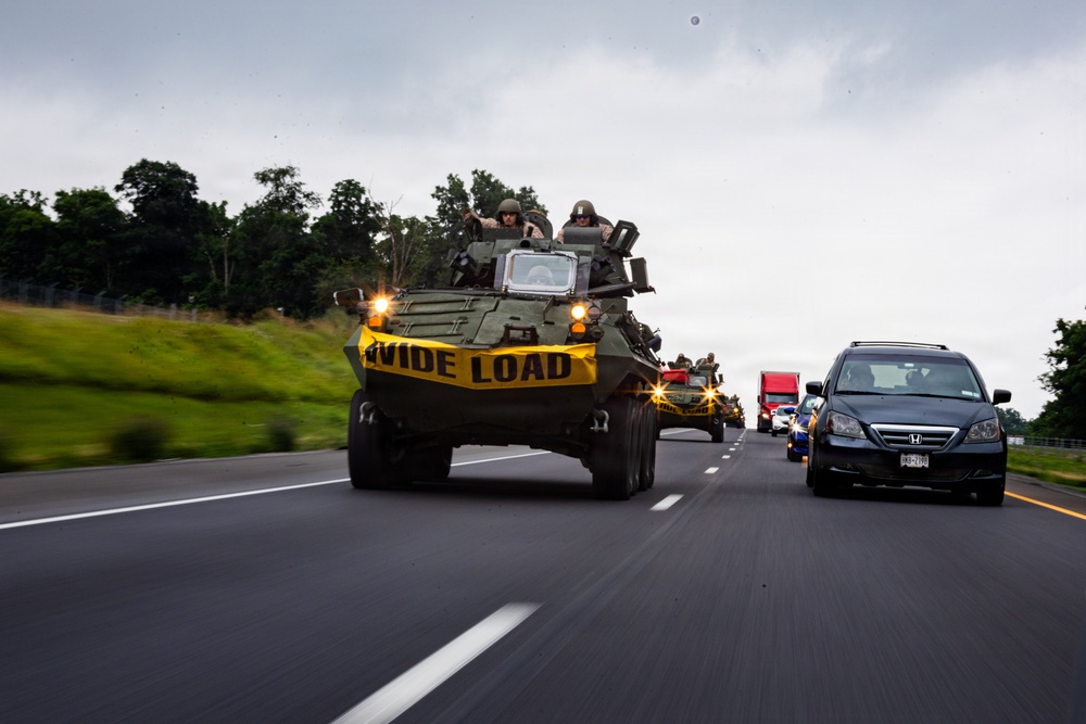 A military armored vehicle convoy is traveling on a public highway alongside civilian traffic. The lead vehicle, an armored personnel carrier with personnel visible in the open turret, displays a yellow 'WIDE LOAD' banner across its front. The convoy occupies the left lane while civilian vehicles, including a Honda Odyssey minivan and a red truck, travel in the right lane.