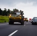 A military armored vehicle convoy is traveling on a public highway alongside civilian traffic. The lead vehicle, an armored personnel carrier with personnel visible in the open turret, displays a yellow 'WIDE LOAD' banner across its front. The convoy occupies the left lane while civilian vehicles, including a Honda Odyssey minivan and a red truck, travel in the right lane.