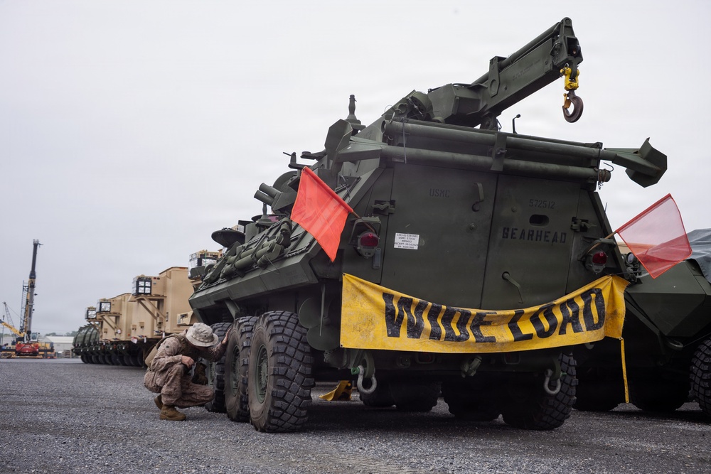 A military recovery vehicle marked 'GEARHEAD' with USMC markings and serial number 572512 is staged on a paved surface, bearing a yellow 'WIDE LOAD' banner and red safety flags on both ends. A service member in camouflage uniform is crouched near the rear undercarriage, apparently inspecting or performing maintenance. The background shows additional military and heavy equipment staged in what appears to be a port or marshaling yard.