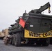A military recovery vehicle marked 'GEARHEAD' with USMC markings and serial number 572512 is staged on a paved surface, bearing a yellow 'WIDE LOAD' banner and red safety flags on both ends. A service member in camouflage uniform is crouched near the rear undercarriage, apparently inspecting or performing maintenance. The background shows additional military and heavy equipment staged in what appears to be a port or marshaling yard.