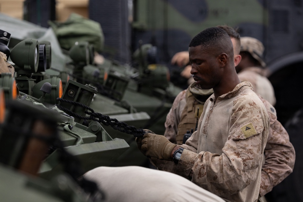 A U.S. Marine in desert MARPAT camouflage uniform is working with a chain attached to a military armored vehicle, likely securing or unsecuring equipment. Additional personnel in military uniforms are visible in the background. The setting appears to be aboard a naval vessel or in a staging area with multiple armored vehicles present.