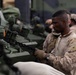A U.S. Marine in desert MARPAT camouflage uniform is working with a chain attached to a military armored vehicle, likely securing or unsecuring equipment. Additional personnel in military uniforms are visible in the background. The setting appears to be aboard a naval vessel or in a staging area with multiple armored vehicles present.
