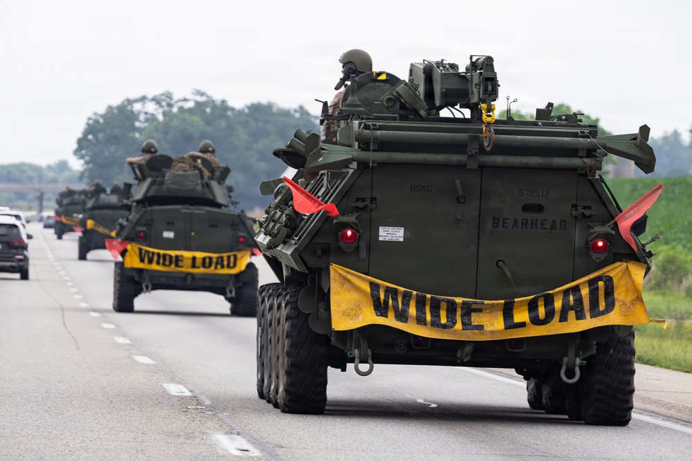 A convoy of USMC (United States Marine Corps) armored vehicles is traveling on a public highway. The lead vehicle is marked 'BEARHEAD' with serial number 572512, and all vehicles display yellow 'WIDE LOAD' banners across their rears. Personnel in military gear are visible in the open hatches atop the vehicles.