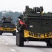 A convoy of USMC (United States Marine Corps) armored vehicles is traveling on a public highway. The lead vehicle is marked 'BEARHEAD' with serial number 572512, and all vehicles display yellow 'WIDE LOAD' banners across their rears. Personnel in military gear are visible in the open hatches atop the vehicles.
