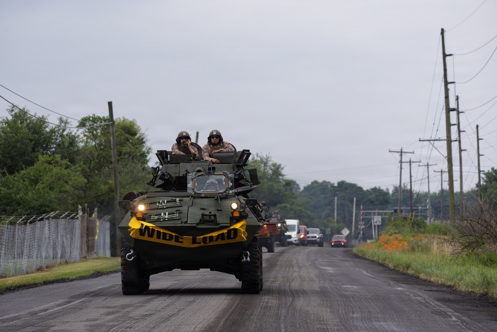 A military armored vehicle, marked with a yellow 'WIDE LOAD' banner, travels along a two-lane rural road with two uniformed personnel visible in the open turret hatch. A convoy of additional military and civilian vehicles follows behind on the same road. The setting appears to be a semi-rural area with utility poles, chain-link fencing, and overcast skies.