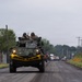 A military armored vehicle, marked with a yellow 'WIDE LOAD' banner, travels along a two-lane rural road with two uniformed personnel visible in the open turret hatch. A convoy of additional military and civilian vehicles follows behind on the same road. The setting appears to be a semi-rural area with utility poles, chain-link fencing, and overcast skies.