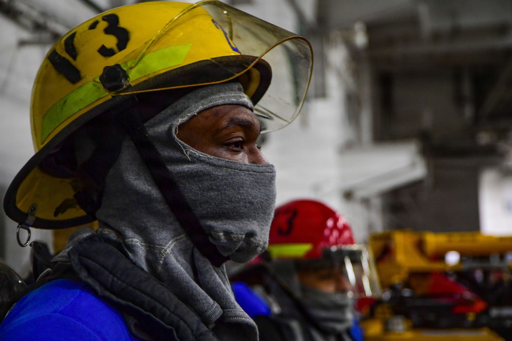 The image shows workers or emergency responders wearing protective helmets with face shields and cloth face coverings wrapped around their heads and faces. The foreground subject wears a yellow helmet marked with the number '3' and a clear visor, along with a gray cloth balaclava-style face wrap. A second person in a red helmet is visible in the background, suggesting a team environment, likely a drill or industrial safety exercise.