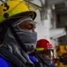 The image shows workers or emergency responders wearing protective helmets with face shields and cloth face coverings wrapped around their heads and faces. The foreground subject wears a yellow helmet marked with the number '3' and a clear visor, along with a gray cloth balaclava-style face wrap. A second person in a red helmet is visible in the background, suggesting a team environment, likely a drill or industrial safety exercise.