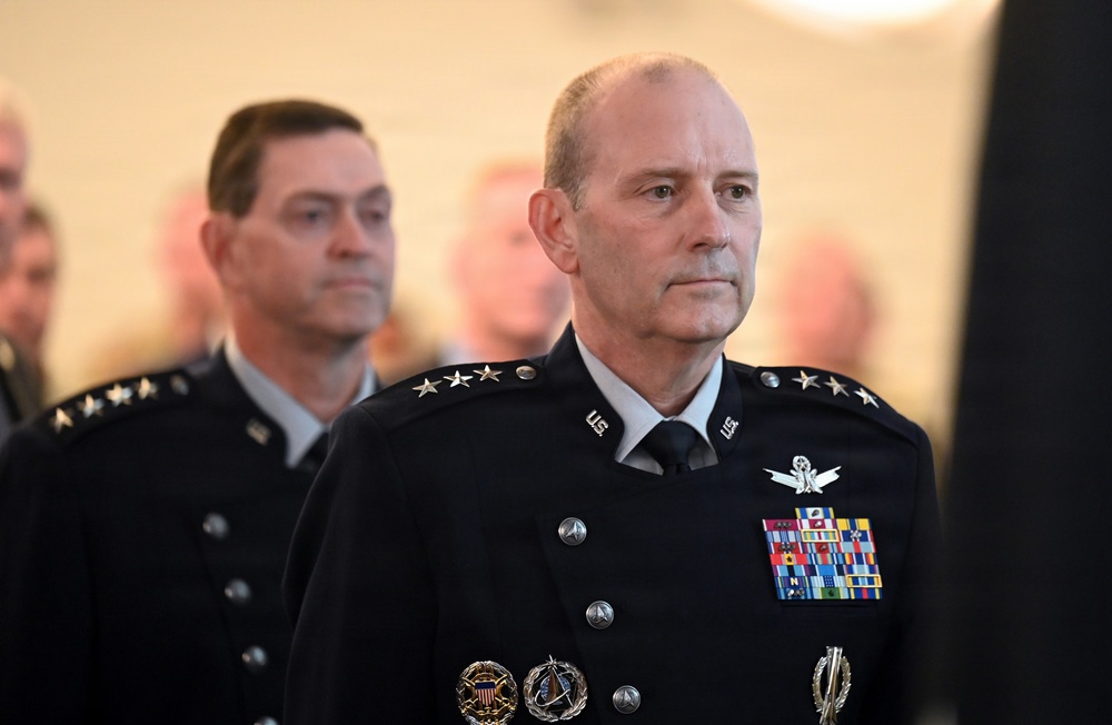 Two senior military officers in dark dress uniforms are visible, with the foreground officer displaying multiple stars on his shoulders indicating high-ranking general officer status. Both wear what appears to be United States Space Force or Air Force dress uniforms with extensive medal ribbons and insignia. The setting appears to be a formal military ceremony or official event.