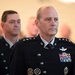 Two senior military officers in dark dress uniforms are visible, with the foreground officer displaying multiple stars on his shoulders indicating high-ranking general officer status. Both wear what appears to be United States Space Force or Air Force dress uniforms with extensive medal ribbons and insignia. The setting appears to be a formal military ceremony or official event.